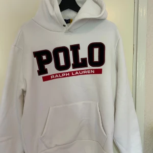 Vit hoodie från Polo Ralph Lauren - Säljer en vit hoodie från Polo Ralph Lauren med stor svart och röd POLO-logga framtill. Tröjan har huva med snörning, magficka och är fodrad med mjukt material. Perfekt för dig som gillar klassisk streetstyle och vill ha något riktigt bekvämt.
