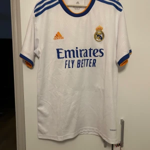 Real Madrid Vini Jr. 20 Adidas tröja - Säljer en officiell Real Madrid fotbollströja från Adidas med Vini Jr. och nummer 20 på ryggen. Tröjan är vit med blå och gula detaljer, kortärmad och tillverkad i lätt funktionsmaterial. Klassisk klubbmärke på bröstet och Emirates-sponsring framtill.