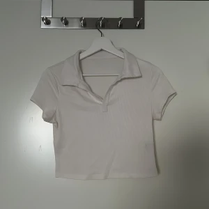 Vit croppad topp med krage - Snygg vit croppad topp med klassisk krage och korta ärmar. Perfekt för en clean och fräsch look, toppen har en enkel design som passar till det mesta. Mjuk och stretchig känsla.