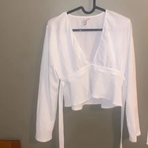 Vit omlottopp med långa ärmar - Säljer en fräsch vit omlottopp med långa ärmar och knytband i midjan där bak. Toppen har en djup v-ringning och är croppap.