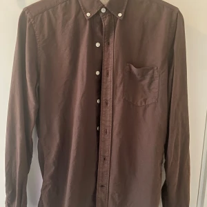 Brun skjorta från A Day's March - Snygg brun skjorta från A Day's March i ekologisk bomull. Skjortan har klassisk krage med knappar, bröstficka och vita knappar framtill. Passformen är normal och ärmarna är långa. Perfekt för dig som gillar stilrena och enkla plagg.