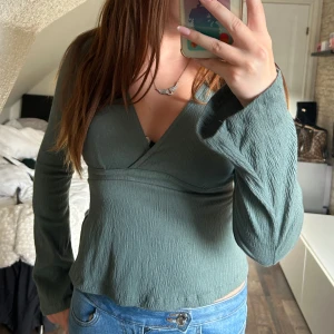 Grön omlottopp med knyt i ryggen - Snygg grön omlottopp med v-ringning och långa ärmar. Toppen har en lätt skrynklig struktur och knyts i ryggen för en extra detalj. Perfekt till jeans eller kjol för en chill och trendig look. Från Gina