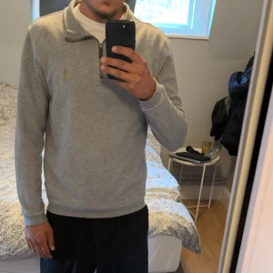 Grå half zip tröja från Polo Ralph Lauren - Snygg grå långärmad tröja från Polo Ralph Lauren med klassisk krage och half zip-dragkedja. Broderad logga på bröstet och mjukt bomullsmaterial. Perfekt till jeans för en clean och stilren look.