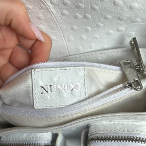Vit axelväska från Núnoo i äkta skinn - Snygg vit axelväska från Núnoo i präglat skinn med cool struktur. Väskan har justerbar axelrem, flera fack med dragkedja och silverfärgade metalldetaljer. Perfekt storlek för det viktigaste och en stilren look till din outfit. Jag fick med dem lapparna när jag köpte vet inte vad som menas men tog med en bild på det ändå.