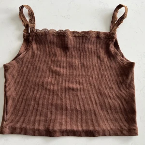 Brun ribbad linnetopp med spets från H&M 134/140 - Säljer en brun ribbad linnetopp från H&M med spetsdetaljer längs axelbanden och upptill. Toppen är ärmlös och har en croppad passform, perfekt för varma dagar.