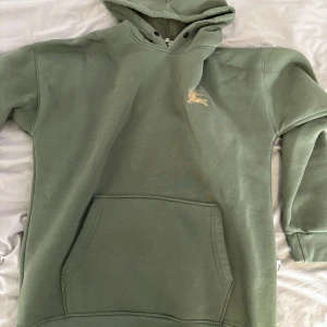 Grön hoodie från Burberry - Säljer en stilren grön hoodie från Burberry med klassisk känguruficka och huva. Tröjan har en liten broderad logga på bröstet och är perfekt för en avslappnad look.
