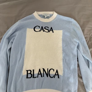 Ljusblå stickad tröja från Casablanca - Säljer en ljusblå stickad tröja från Casablanca med vit panel framtill och svart text 'CASA BLANCA'. Tröjan har rund halsringning och ribbade muddar i vitt. Perfekt för dig som gillar stilrena och trendiga plagg.