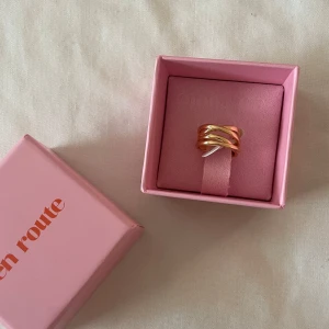 Guldfärgad ring från En Route - Aldrig använd! Trendig guldfärgad ring från En Route med vågig, flätad design. Ringen levereras i en söt rosa ask och passar perfekt som statement-accessoar. Snygg att bära ensam eller att kombinera med andra ringar.