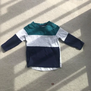 Randig långärmad tröja med knappar på axeln - Säljer en långärmad tröja i bomull med breda blockränder i mörkgrönt, ljusgrått och marinblått. Tröjan har praktiska tryckknappar på ena axeln för enkel påklädning. Perfekt för en avslappnad och stilren look.