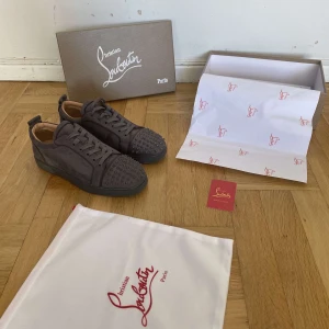 Gråa sneakers från Christian Louboutin med nitar - Säljer ett par gråa sneakers från Christian Louboutin med matta nitar framtill och klassisk röd sula. Skorna är i mocka och har snörning. Kommer med originalkartong och dustbag.