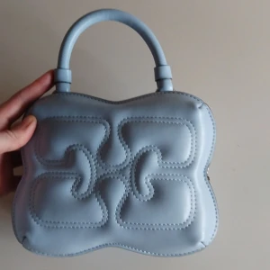 Ganni väska  - Säljer en ljusblå handväska med unik vadderad design och matchande avtagbar axelrem. Väskan har ett handtag upptill och stängs med dragkedja. Perfekt för dig som vill ha en snygg accessoar med cool form och detaljer.