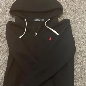 Svart hoodie från Polo Ralph Lauren - Svart hoodie från Polo Ralph Lauren med vit dragsko och dragkedja. Klassisk röd broderad logga på bröstet. Perfekt för en avslappnad och stilren look. Fickan var sönder men sydde den.