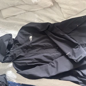 Svart vindjacka från The North Face - Säljer en svart vindjacka från The North Face med huva och dragkedja framtill. Jackan har justerbara ärmslut med kardborre och logga på bröstet. Perfekt för blåsiga dagar och enkel att matcha med olika outfits😊