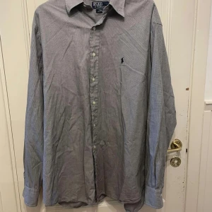 Rutig skjorta från Polo Ralph Lauren - Klassisk svartvit rutig skjorta från Polo Ralph Lauren i 100% bomull. Skjortan har långa ärmar, knappar framtill och den ikoniska broderade loggan på bröstet. Perfekt för en stilren och tidlös look.