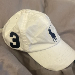 Vit keps från Polo Ralph Lauren - Vit keps från Polo Ralph Lauren med mörkblå broderad logga och siffra 3 på sidan. Helt ny och aldrig använd