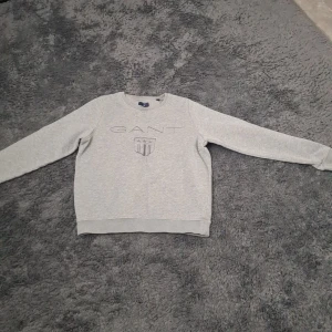 Grå sweatshirt från GANT - Säljer en stilren grå sweatshirt från GANT med broderad logga och sköld på bröstet. Tröjan har rund halsringning och långa ärmar. Perfekt för en avslappnad och klassisk look.