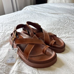 Bruna sandaler med spänne - Snygga bruna sandaler med breda korsade remmar och ett stort guldfärgat spänne vid ankeln. Skorna har platt sula och öppen tå, perfekta för varma dagar. Tillverkade i skinnliknande material för en stilren look. Prislappen är kvar!