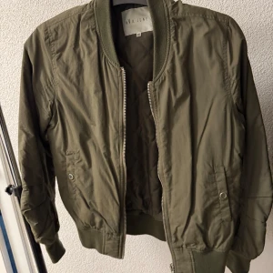 Olivgrön bomberjacka  - Säljer en olivgrön bomberjacka. Jackan har klassisk bombermodell med ribbade muddar, dragkedja framtill och fickor med tryckknapp. Perfekt för vår och höst. Snygg och enkel att matcha till olika outfits.