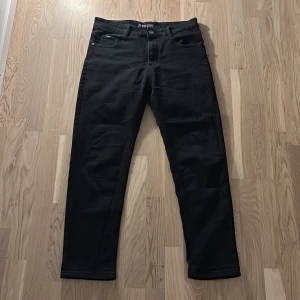 Svarta jeansbyxor från Fashion - Svarta jeansbyxor från Fashion med klassisk femficksdesign och raka ben. Byxorna har knappgylf och bälteshällor, samt Fashion-logga på bakfickan och läderpatch i midjan. Perfekta för en stilren och enkel look.