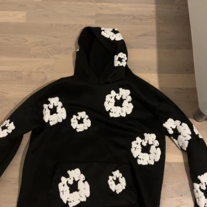 Svart hoodie med vita blommor från Denim Tears - Säljer en svart hoodie från Denim Tears med stora vita blommor tryckta över hela tröjan. Hoodien har en klassisk passform med huva och magficka. Perfekt för dig som vill sticka ut med en unik och trendig stil.