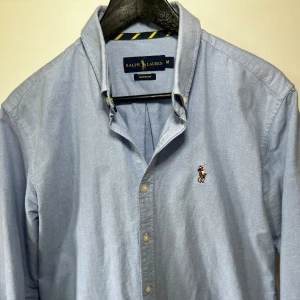 Ljusblå skjorta från Ralph Lauren - Klassisk ljusblå skjorta från Ralph Lauren. Skjortanär i bomull och är custom fit M. Skick 10/10 typ avvänd nån enstaka gång. Köpt för 1600kr och säljer för 500kr.