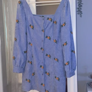 Blå klänning med broderade blommor och knappar - Supersöt blå klänning med broderade gula blommor och gröna blad. Klänningen har långa ärmar med knappar vid ärmslut och framtill. Den har en fyrkantig urringning och är i ett mjukt, lite ribbat material. Perfekt för vår och sommar!