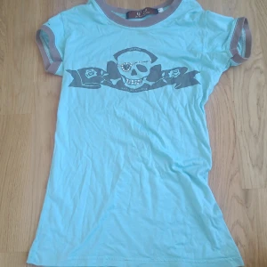 Vintage 2000-stils topp - Cool ljusblå t-shirt från G'29 med gråa muddar och ett stort döskalletryck framtill. Trycket har detaljer med nitar på ögonlapp och tänder. Perfekt för dig som gillar edgy stil!