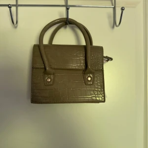 Beige mini handväska med krokodilpräglat mönster - Säljer en beige mini handväska med krokodilpräglat mönster och två handtag. Väskan har även en lång axelrem som går att ta av. Perfekt för dig som vill ha en stilren accessoar med trendig känsla.