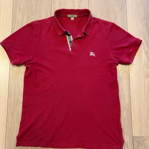 Röd pikétröja från Burberry  - Säljer en klassisk röd pikétröja från Burberry Brit med korta ärmar och ikonisk rutdetalj vid knäppningen. Tröjan har broderad logga på bröstet och snygg krage. Perfekt för dig som gillar stilrena märkeskläder. Väldigt bra skick. 