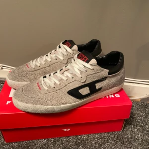 Grå sneakers från Diesel - Säljer ett par gråa sneakers från Diesel med svart logga på sidan och svarta detaljer vid hälen. Skorna har vit sula med röd undersida och vita snören. Diesel-loggan syns tydligt på plösen. Perfekta för dig som gillar streetstyle.