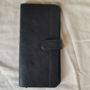 Svart plånbok i skinn - Säljer en stilren svart plånbok i skinn med flera kortfack och dragkedjefack. Plånboken har en enkel design med tryckknappsstängning och gott om plats för både kort och sedlar. Perfekt för dig som vill ha en klassisk och praktisk accessoar.