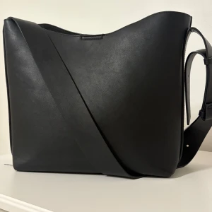 Svart axelväska i skinn - Säljer en stilren svart axelväska i skinn med bred axelrem. Rymmer mycket, inge fack på insidan. Lätt att matcha med olika outfits. Pris kan justeras!