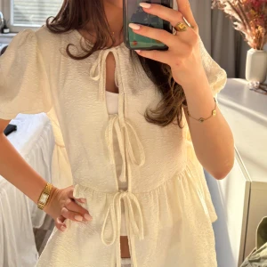 Vit blus med knytband - Söt vit blus med puffärmar och tre knytband framtill. Blusen har en lätt struktur i tyget och en lös passform som ger en romantisk känsla. Perfekt att styla med jeans eller kjol för en fräsch look.