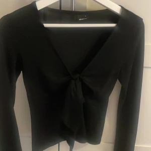 Svart långärmad topp med knyt från Gina Tricot - Snygg svart långärmad topp från Gina Tricot med v-ringning och knyt framtill. Perfekt att styla med jeans eller kjol för en trendig look. Mjuk och stretchig känsla.