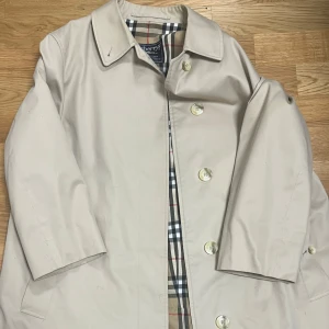 Beige trenchcoat från Burberrys - Klassisk beige trenchcoat från Burberrys med ikoniskt rutigt foder i beige, svart, vitt och rött. Jackan har knappar framtill och en tidlös krage. Perfekt för dig som gillar stilrena och tidlösa plagg.