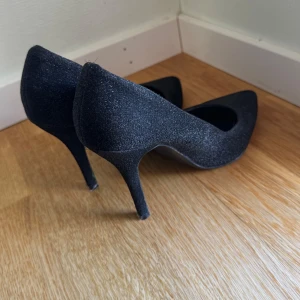 Svarta glittriga pumps från Bershka - Säljer ett par svarta pumps från Bershka med glittrigt material och spetsig tå. Skorna har hög klack och en elegant form som passar perfekt till festliga tillfällen. Klassisk och stilren design med platt sula undertill.