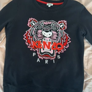 Svart sweatshirt med tigerbrodyr från Kenzo - Svart sweatshirt från Kenzo med ikonisk tigerbrodyr och texten 'KENZO PARIS' framtill. Tröjan har rund halsringning, långa ärmar och ribbade muddar. Broderiet har detaljer i rött, vitt och rosa. Perfekt statement-plagg för din garderob. Den har ett litet hål på vänster arm (bild 5) men inget som syns och om man vill enkelt att sy ihop
