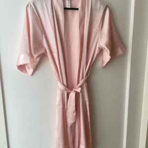 Kimono från Missäy - Säljer en ljusrosa kimono från Missäy i satinliknande material. Klänningen har korta ärmar, knytband i midjan och en öppen front. Perfekt för en mjuk och elegant look! Enbart provad på och har inga defekter ☺️