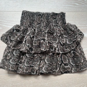 Brun volangkjol med ormmönster - Säljer en kort volangkjol med trendigt ormmönster i bruna och svarta toner. Kjolen har bred resår i midjan och dubbla volanger som ger en snygg och livlig look. 