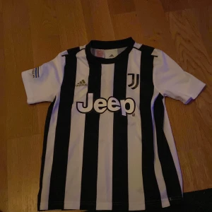 Juventus fotbollströja med svarta och vita ränder - Juventus matchtröja från Adidas med klassiska svarta och vita vertikala ränder. Jeep-logga på bröstet och klubbmärke på vänster sida. Kortärmad modell med rund hals och Adidas-logga på höger sida.