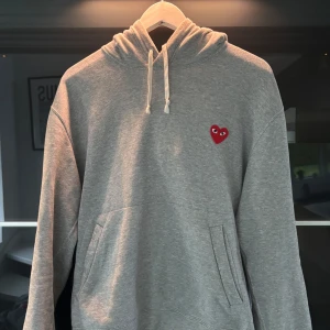 Grå hoodie från Comme des Garçons Play - Grå hoodie från Comme des Garçons Play med klassisk huva och dragsko. På bröstet finns det ikoniska röda hjärtat med ögon. Tröjan har känguruficka framtill och är tillverkad i mjuk bomull. Storlek m men passar S