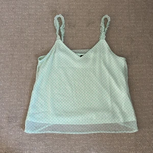 Ljusgrön prickig linnetopp från Vero Moda - Söt ljusgrön linnetopp från Vero Moda med små mintgröna prickar och tunna, rynkade axelband. Toppen har v-ringning och ett lätt transparent ytterlager som ger en luftig känsla. Perfekt för varma dagar! (Säljer i brist på användning)