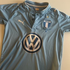 Malmö FF fotbollströja från Puma - Ljusblå Malmö FF fotbollströja med klubbmärke, guldstjärna och stor Volkswagen-logga på bröstet. Tröjan har korta ärmar, knappslå vid halsen och vita Puma-detaljer. Perfekt för dig som vill visa ditt stöd för MFF. Tröjan är i storlek 140
