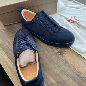 Mörkblå sneakers från Christian Louboutin - Säljer ett par mörkblå sneakers från Christian Louboutin med klassisk röd sula och diskret broderad logga på hälen. Skorna har snörning och är tillverkade i mocka med beige innersida. Perfekta för dig som vill sticka ut med stil.