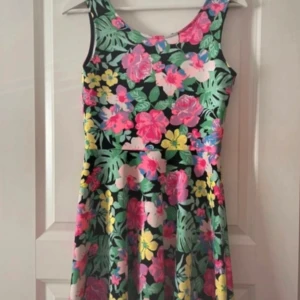 Blommig klänning - Säljer en blommig klänning med rund halsringning och ärmlös design. Klänningen har en utställd kjol och mönster i rosa, gul, grön och blå. Perfekt för varma dagar och somriga tillfällen!