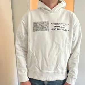 Vit hoodie med tryck från Acne Studios - Säljer en vit hoodie från Acne Studios med svart tryck och världskarta på bröstet. Texten 'PAOUASIE NOUVELLE GUINEE' och fransk text ovanför. Klassisk huva och normal passform. Perfekt för en stilren och trendig look.