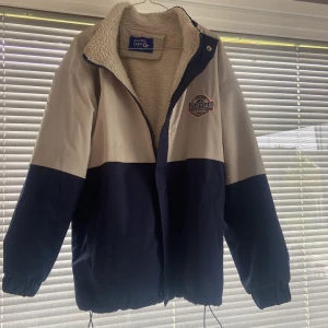 Blå och vit teddyfodrad jacka från Aelfric Eden - Blå och vit jacka från Aelfric Eden med teddyfoder. Jackan har dragkedja, hög krage och broderad patch på bröstet. Har justerbar dragsko nertill och är oversized i storleken. Säljer p.g.a för stor.