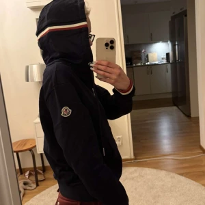 Svart hoodiejacka från Moncler - Mörkblå hoodie från Moncler med dragkedja framtill och huva. Hoodien har vita och röda ränder på huvan och ärmsluten, samt Moncler-logga på ärmen. Perfekt för en sportig och avslappnad stil. Passar till alla och är så mysig! Helt oanvänd endast tagit bilder i tröjan!