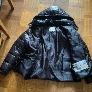 Svart dunjacka från Moncler - Säljer en svart dunjacka från Moncler med glansig finish och klassisk Moncler-logga på ärmen. Jackan har huva, dragkedja och stora fickor med lock. Perfekt för kyliga dagar och riktigt snygg streetstil.
