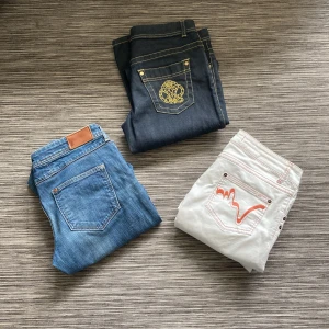 Tre par jeans i olika färger och stilar - Säljer tre par jeans: ett par mörkblå med gul brodyr från H&M x Roberts Cavalli, ett par klassiska blå Flary jeans och ett par vita jeans från Ichi med röda sömmar. Perfekta för dig som vill variera din stil med olika färger och detaljer| Storleken på Jensen ser du på bilderna| Hör av dig vid fundering 🤗